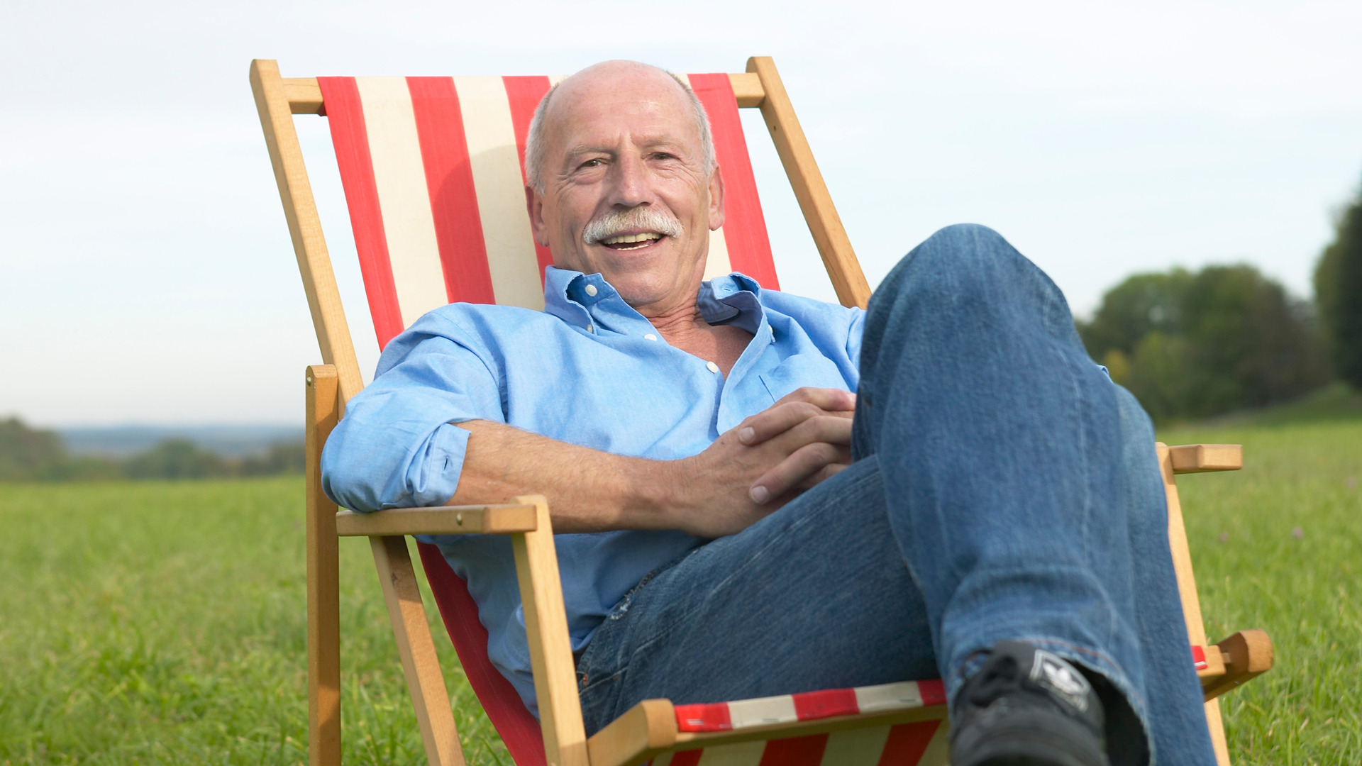 Eldre mann med bart og uten hår sitter i en solstol med røde og hvite striper